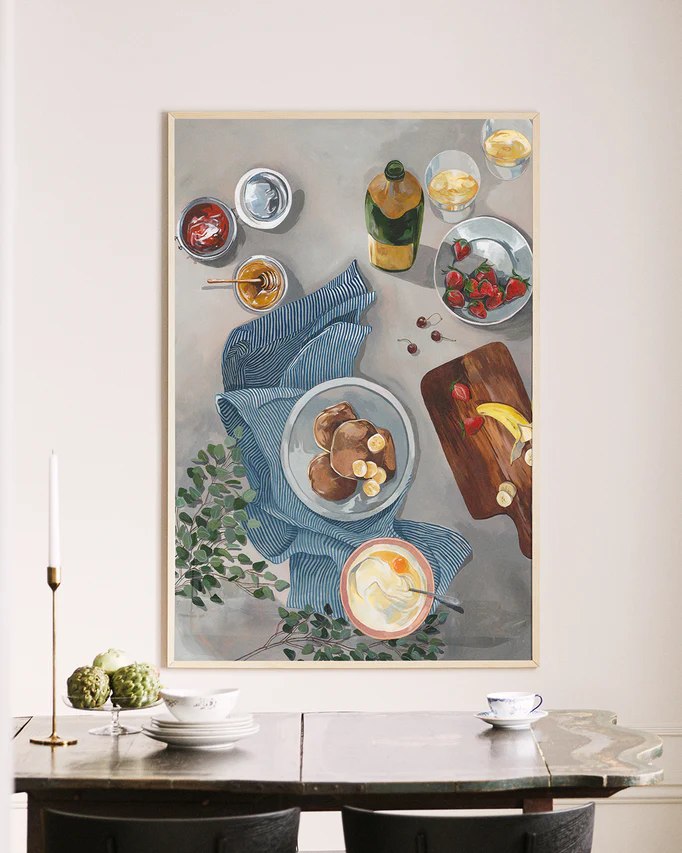The wall features a Breakfast of Champions Poster showcasing a delightful breakfast scene with fluffy pancakes, fruit, drinks, a striped cloth, bananas and strawberries on a cutting board, and a bottle. Below the dining table are chairs, napkins, and a candle.