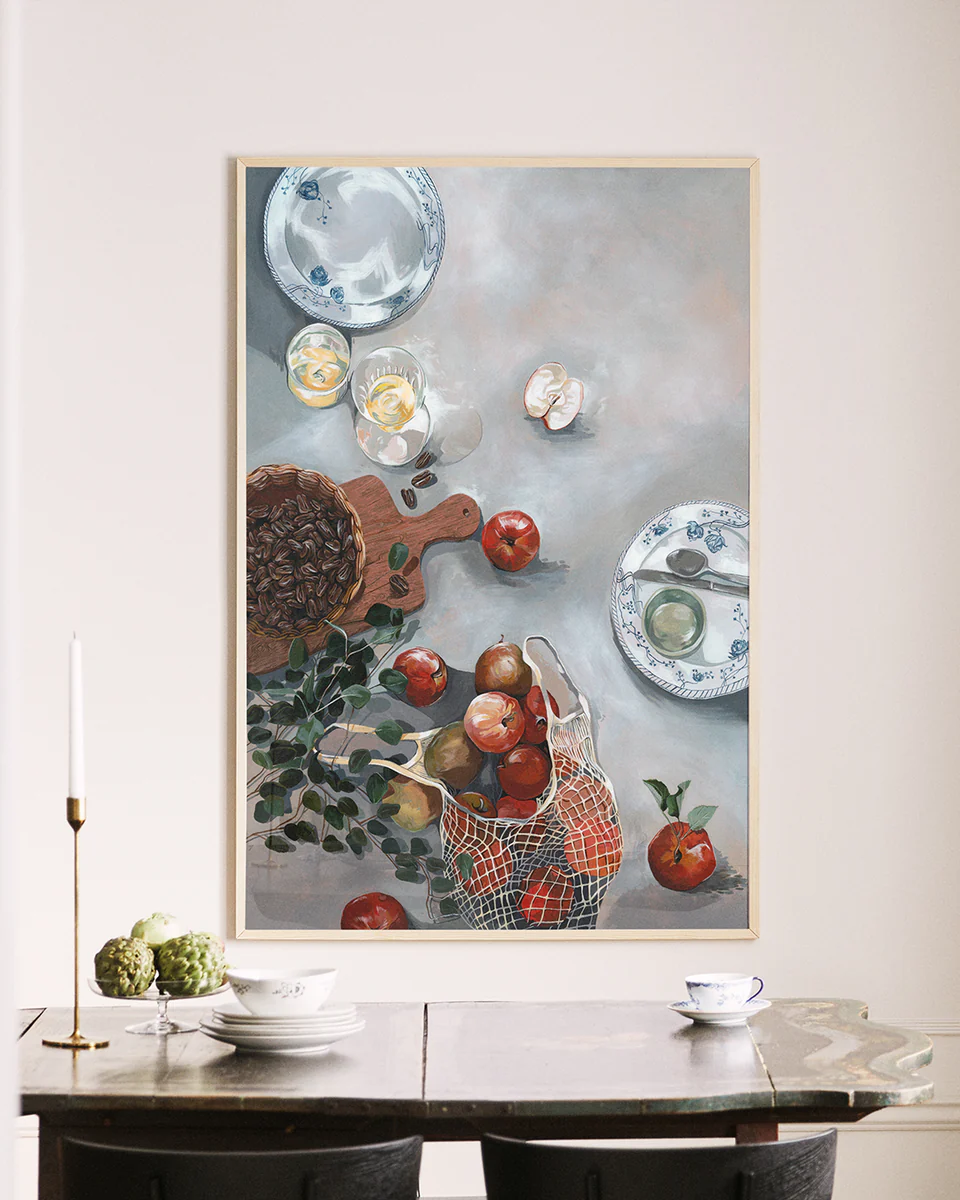 The How Do You Like These Apples poster, showcasing a table with red apples, a basket, and dishes, hangs on the wall. Below it, a kitchen table with a candlestick, small plant, and two dark chairs seamlessly blends this art piece with reality through its warm decor.