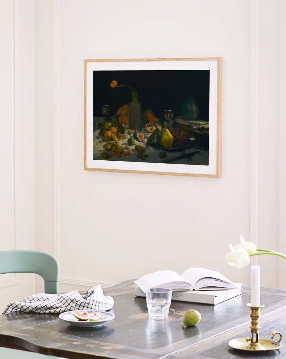 An open book, glass of water, and candlestick create a tranquil dining table scene with a single tulip in a vase nearby. A framed Gloomy Picnic II Poster reminiscent of Paul Cézannes style adorns the light-colored wall.