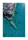 A small woman swimming alone in a big blue sea next to a grey cliff.