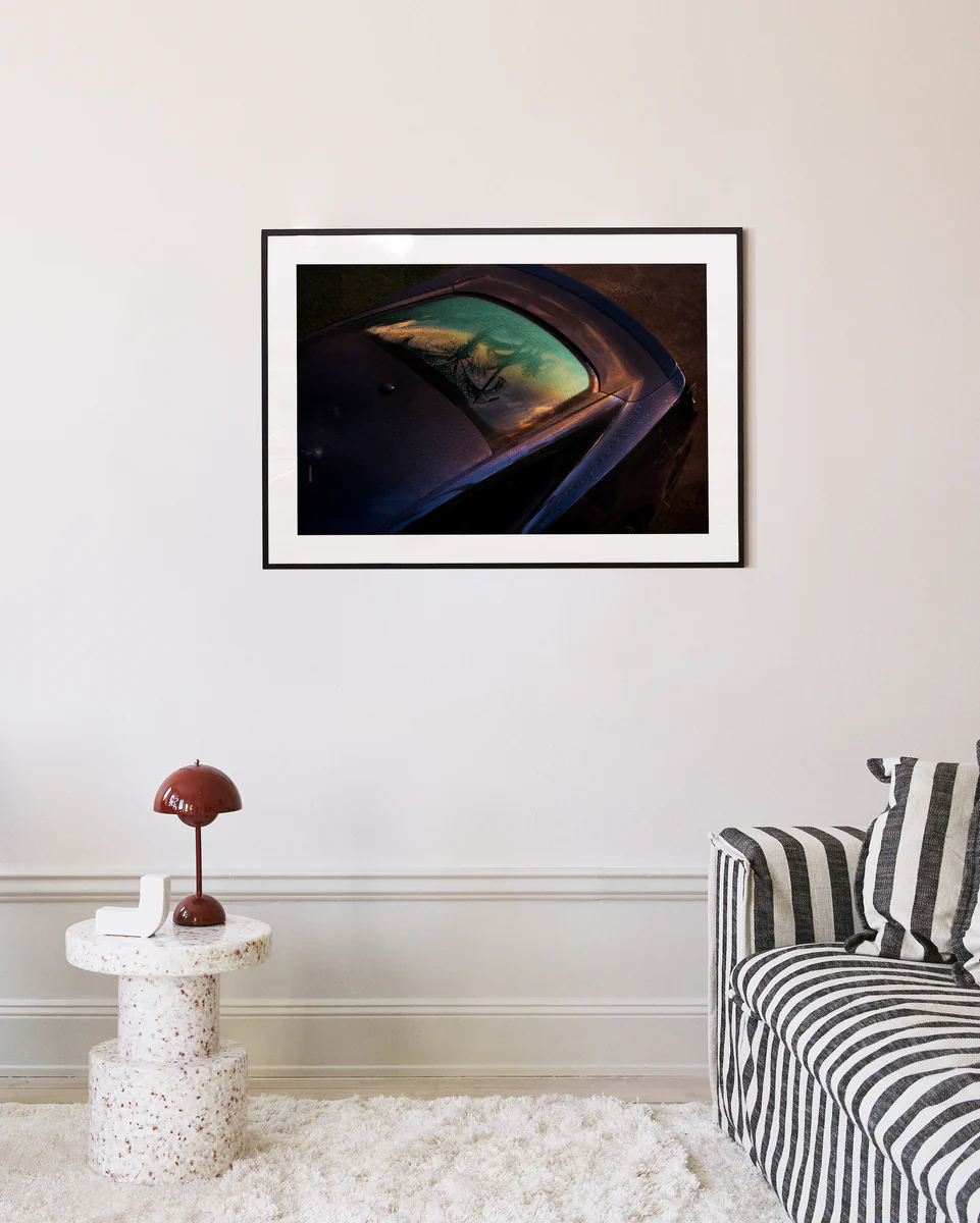 A modern living room boasts a striped sofa, a round white speckled side table with a brown lamp, and the Gas Up Poster, capturing the pedal to the metal spirit with car reflections of trees and sky on the cream-colored wall.