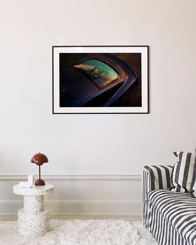 A modern living room boasts a striped sofa, a round white speckled side table with a brown lamp, and the Gas Up Poster, capturing the pedal to the metal spirit with car reflections of trees and sky on the cream-colored wall.