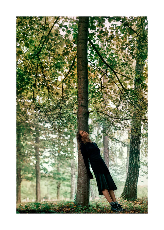 Woman leaning against a tree trunk in the forest and looking up into its crown.