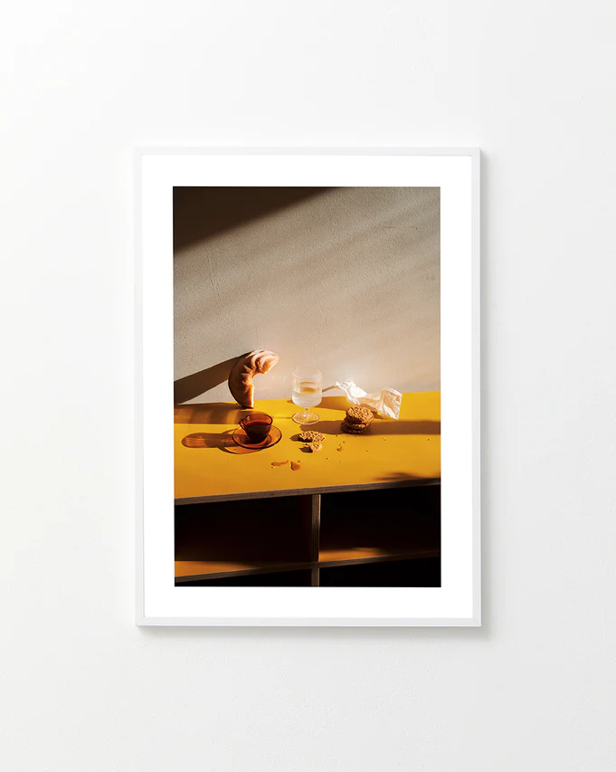 The Would You Like Some Coffee? poster depicts a still life on a white wall featuring a croissant, glass of water, wooden bowl, and cookies on a yellow shelf bathed in sunlight, with long shadows cast across the scene.