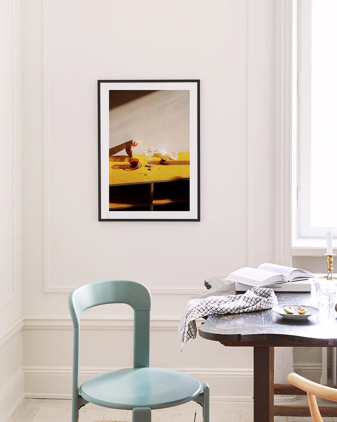 A minimalist room features a Would You Like Some Coffee? poster on a white wall, showing a hand pouring liquid into a cup on a yellow table. Nearby are a blue chair and dark wooden table with an open book and patterned cloth, all bathed in natural light.
